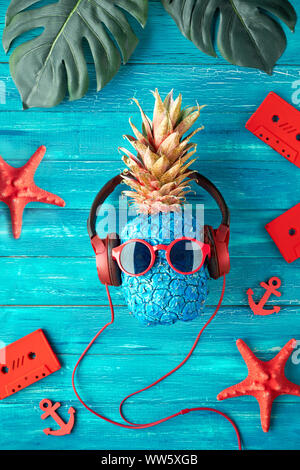 Tanga carattere di ananas in rosso gli occhiali da sole e auricolari ascolta musica retrò. Testurizzato rustico in legno pannelli sfondo dipinto di blu, vista dall'alto, f Foto Stock