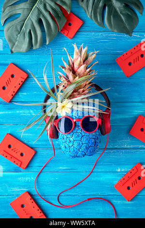 Tanga carattere di ananas in rosso gli occhiali da sole e auricolari ascolta la musica. Rustico in legno a trama sfondo blu con red cassette audio e Foto Stock