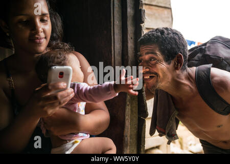 Tre generazioni, giovane donna seduta con il suo bambino e utilizzando il suo telefono cellulare, il nonno di fare divertire con il bambino sul suo braccio Foto Stock