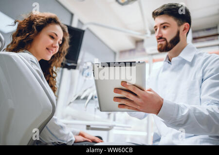 Felice dentista e paziente commentando trattamenti in un tablet applicazione in una consultazione con attrezzature mediche in background. Foto Stock