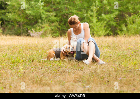 Donna attraente in un top bianco e jeans blu seduta su erba, abbracciando gatto bianco, guardando il suo cane, posa vicino a. Foresta verde sullo sfondo Foto Stock