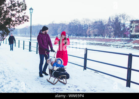 Coppia giovane spingendo la slitta di legno con i loro due bambini piccoli in caldo abbigliamento invernale. Bella inverno terrapieno su uno sfondo Foto Stock