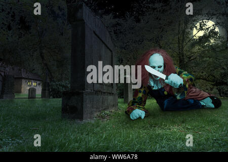 Un pauroso clown con un coltello su un cimitero Foto Stock