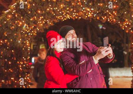 Chiudere la vista ritratto della coppia giovane rendendo selfie Near Fir Tree arch, decorata con ghirlande. Coppia sposata a fotografare durante la vacanza invernale trave Foto Stock