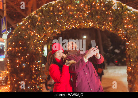 Giovane coppia romantica rendendo selfie vicino decorate arch fatto di verde abete e decorate con luci Foto Stock