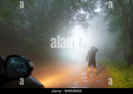 Un science fiction concetto di un uomo che guarda gli alieni che esce la nebbia in una nebbiosa, spooky strada forestale in serata. Evidenziato in auto fari. Foto Stock