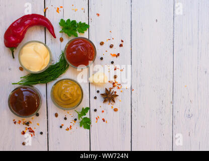 Un set di salse e uno speciale - ketchup, maionese e senape su una piastra bianca, peperoncino, prezzemolo e l'aneto. Vista dall'alto in bianco sullo sfondo di legno. Foto Stock