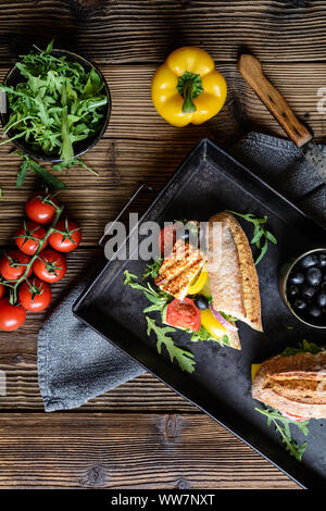 Vegetariani integrali baguette farcite con grigliate di formaggio bianco, rucola, pomodori, olive nere, il peperone e la cipolla rossa su sfondo di legno Foto Stock