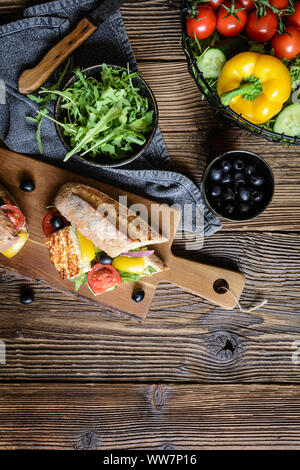 Vegetariani integrali baguette farcite con grigliate di formaggio bianco, rucola, pomodori, olive nere, il peperone e la cipolla rossa su sfondo di legno Foto Stock