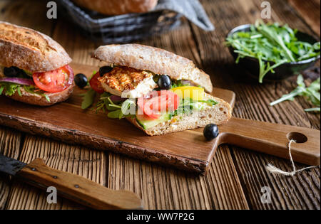 Vegetariani integrali baguette farcite con grigliate di formaggio bianco, rucola, pomodori, olive nere, il peperone e la cipolla rossa su sfondo di legno Foto Stock