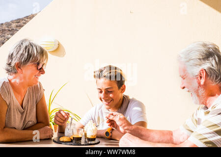 Tre persone un uomo una donna un ragazzo con bel tempo con sorrisi per esterno a casa. bere il caffè con panna e alcuni biscotti. bella la vita di giorno lifestyle con sorrisi e ridere sotto mar luce solare in vacanza Foto Stock