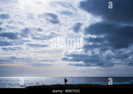 Lonely surfer in silhouette stand up in attesa di qualche le onde dello oceano. toni blu con bellissimo cielo di nuvole sopra di lui. concetto hero Foto Stock