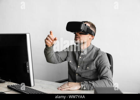 Giovane uomo barbuto seduto contro computer con occhiali per realtà virtuale cercando di toccare qualcosa o toccare qualche cosa di fronte a ciò che egli ha visto. Moderno Foto Stock