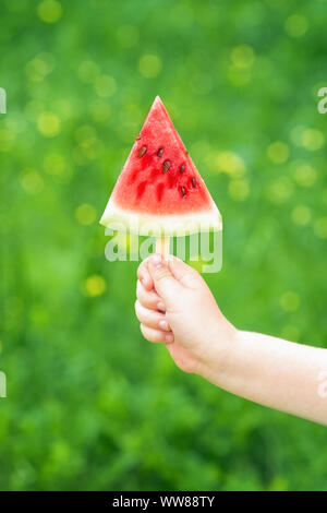 Un bambino la mano è in possesso di una fetta triangolare di un cocomero su un verde natura sfondo, concetto di berry ice cream Foto Stock