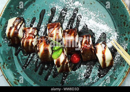 Il dessert di frittelle sotto forma di rotoli con crema di latte ripieno di formaggio con menta su una piastra di blu, con sciroppo e cioccolato topping. Vista da sopra. Foto Stock