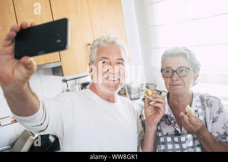 Senior caucasico adulto felice coppia a casa nella cucina cucina e lavorare insieme con gioia e felicità, tenendo selfie con moderne smart phone e mangiare un pezzo di torta, di amore e di vita per sempre concetto Foto Stock