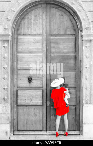 Una donna in un vestito rosso che tiene un bambino, in attesa di qualcuno per aprire la porta grande e lasciarli dentro; Granada, Spagna. Foto Stock