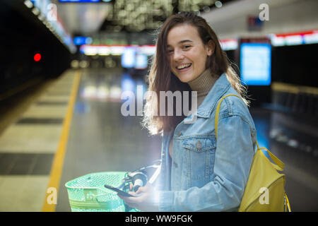 Ragazza adolescente in jeans con zaino giallo e bike permanente sulla stazione della metropolitana, in attesa del treno, sorridere e ridere. Futuristica stazione della metropolitana Foto Stock