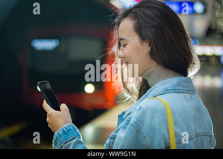 Ragazza adolescente in jeans con zaino permanente sulla stazione metropolitana smart tenendo il telefono in mano, scorrimento e texting, sorridere e ridere. Finlandia Foto Stock