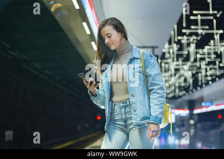 Ragazza adolescente in jeans con zaino permanente sulla stazione metropolitana smart tenendo il telefono in mano, scorrimento e texting, sorridere e ridere. Finlandia Foto Stock