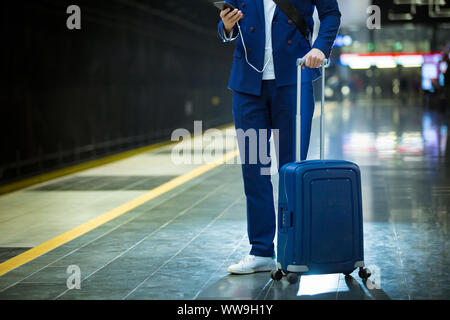 Giovane elegante uomo bello in tuta con valigia permanente sulla stazione metropolitana smart tenendo il telefono in mano, scorrimento e texting, sorridere e ridere. Foto Stock