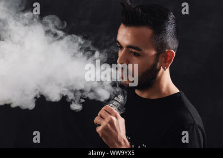 Giovane uomo con la barba vaping sigaretta elettronica su sfondo nero. Foto Stock