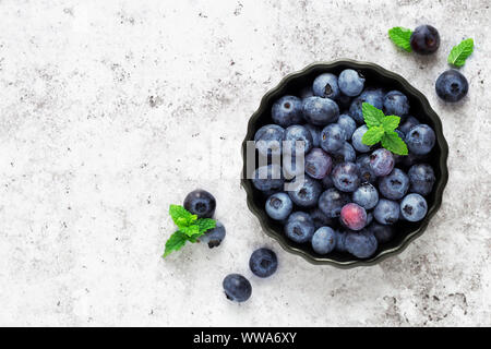 Fresche e mature di mirtilli giardino con ciuffi di menta in una cottura nero ciotola contro il cemento grigio Sfondo. Vista aerea, laici piatta Foto Stock