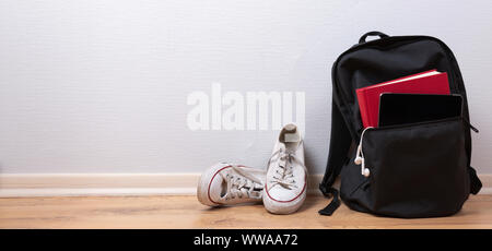 Vecchio sneakers bianco, libro e tablet con auricolari in uno zaino su sfondo di legno. Scuola studente e concetto di viaggio Foto Stock