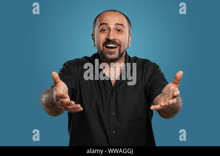 Close-up verticale di un brunet uomo di mezza età con la barba, vestito di una t-shirt nera e in posa contro uno sfondo blu. Foto Stock