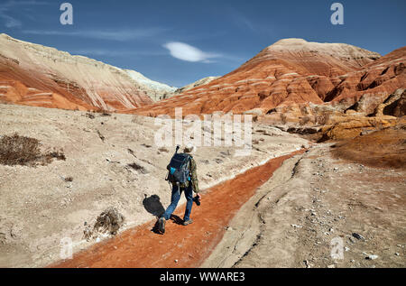 Turistico con zaino e fotocamera a piedi il polveroso canyon su surreale monti rossi contro il cielo blu nel deserto Foto Stock