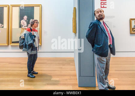 Un vecchio femmina con il suo tour audio telefoni di testa si erge davanti a Francis Bacon trittico in un museo mostra il suo lavoro mentre una parete separa Foto Stock