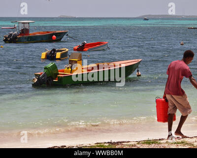 Uomo in una t-shirt rossa e portante un rosso in plastica tanica di benzina sulla spiaggia, con varie barche in background. Island Harbour, Anguilla BWI. Foto Stock