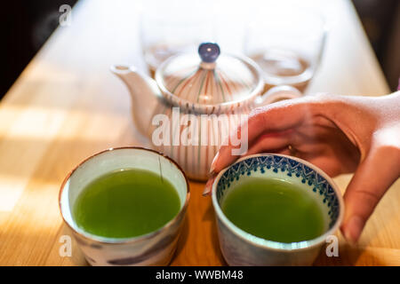 Donna mano closeup holding teacups o tazze di tè da teiera con caldo Giapponese tradizionale Sencha tè verde a cucina tavolo in legno Foto Stock