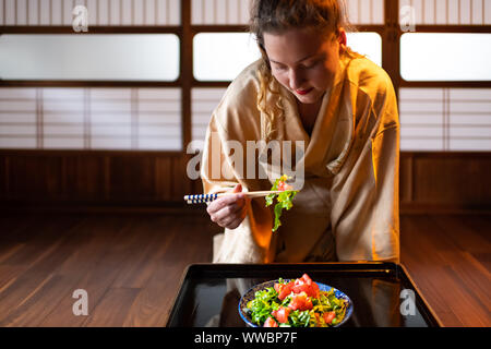 Giovane donna in kimono seiza seduti a casa tradizionale giapponese o ryokan camera da tavola bacchette di contenimento, mangiare insalata dalla piastra di scorrimento shoji Foto Stock