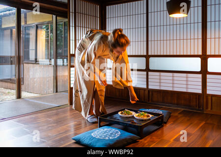 Casa Tradizionale Giapponese o ryokan camera con giovani straniero donna che serve fare soffriggere le verdure di negi, funghi e tofu tabella piastre in kimono wi Foto Stock