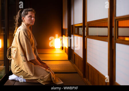 Tradizionale casa giapponese o ryokan e gaijin giovane donna in kimono, dei tabi calzini seiza seduti sul tatami pavimento da porte scorrevoli di carta shoji Foto Stock