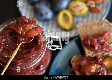 Chutney di prugne o inceppamento in un vasetto di vetro con coperchio aperto e il cucchiaio d'oro nero sul piano portapaziente. Fette di pane integrale e la piastra con le prugne di sfondo sfocato. Foto Stock