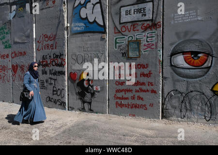 Una donna Palestinese passeggiate lungo la barriera di separazione o parete spruzzata con graffiti di slogan da turisti alla periferia della città di Betlemme in central West Bank. Territori palestinesi, Israele Foto Stock