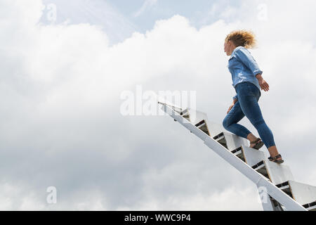 Bionda donna caucasica camminando su per le scale fino al cielo con le nuvole sullo sfondo. Foto Stock