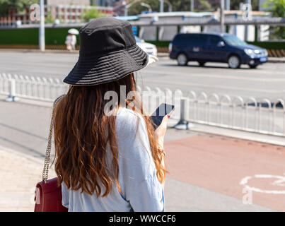 Una donna cinese in un cappello per il sole sorge sul marciapiede e mantiene uno smartphone. Foto ritratto da dietro, volto non è visibile e sfondo sfocato Foto Stock