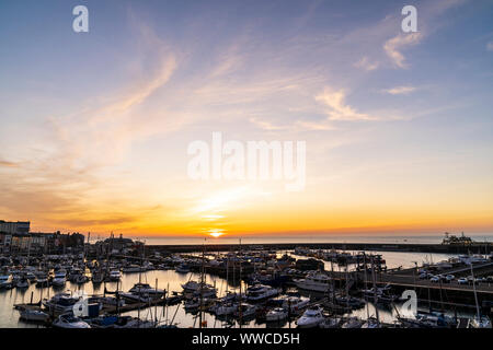 Alba sul Canale della Manica e il porto della città costiera di Ramsgate su alla costa del Kent in Inghilterra. Porto e Marina yachting con una sottile fascia off giallo arancio all'orizzonte con il sole che sorge dal mare, cielo blu con nuvole wispy sopra. Foto Stock