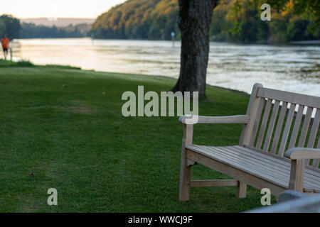 Due panche di legno in angolo di novanta gradi in primo piano sull'erba verde con alberi e passeggiate persona presso la riva di un fiume svizzero Rhein al tramonto. Foto Stock