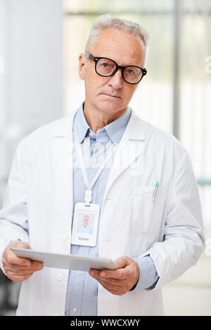 Vita ritratto di senior medico guardando la telecamera durante l'uso di digitale compressa in piedi in ufficio di moderna clinica Foto Stock
