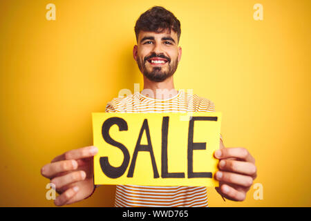 Giovane uomo con tatuaggio azienda vendita banner in piedi isolato su sfondo giallo con una faccia felice in piedi e sorridente con un sorriso sicuro showi Foto Stock