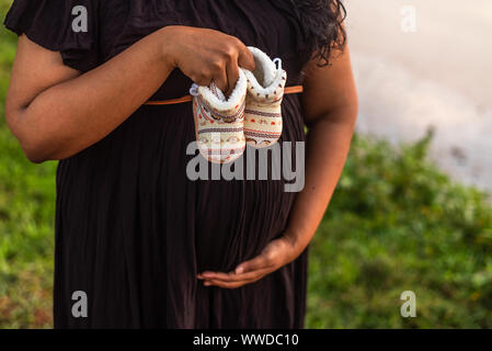 Donna incinta in abito nero azienda baby stivali e visualizza il suo bambino bump in corrispondenza di un outdoor shoot Foto Stock