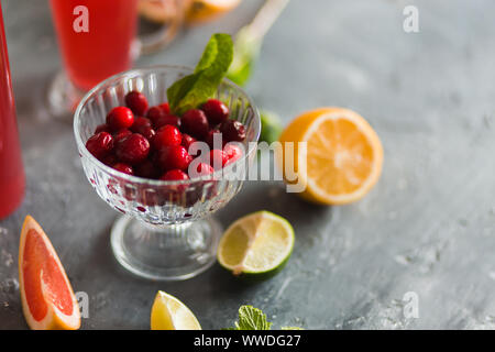 Vista dettagliata del rosso di mirtilli rossi maturi in una ciotola con agrumi e bevande su uno sfondo grigio. Bacche e frutti, vitamine, bibite estive concept Foto Stock