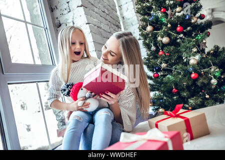 Allegra donna felice con la figlia seduta sul pavimento nel soggiorno vicino a albero di natale. Nuovo Anno di interni Foto Stock