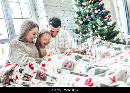 Abbracciando la famiglia vicino albero di natale sul letto. Nuovo anno di mattina. Accogliente vacanza all'abete. amore, felicità e il concetto di famiglia Foto Stock