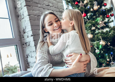 Piccola ragazza baciare la sua mamma sulla guancia su albero di Natale sfondo. Trascorrere Capodanno a casa insieme, happy family concept