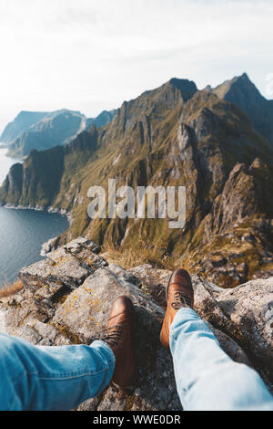 Viaggiatore Da Solo seduto sul bordo della montagna sopra il mare. POV vista sulle grandi montagne e scarponcini da trekking. Foto Stock
